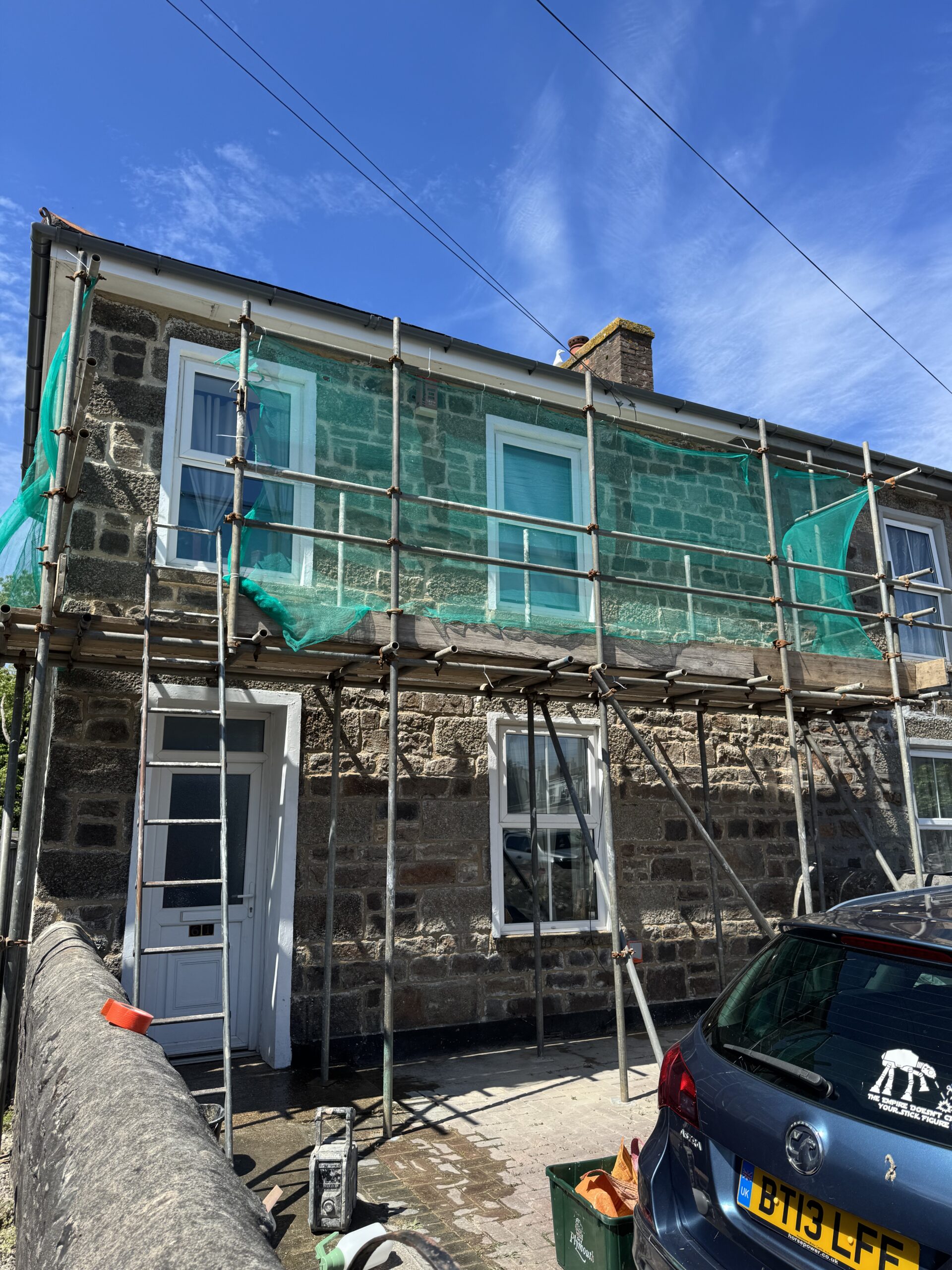 A traditional two-story stone house covered in construction scaffolding with green safety netting under a blue sky in Cornwall.