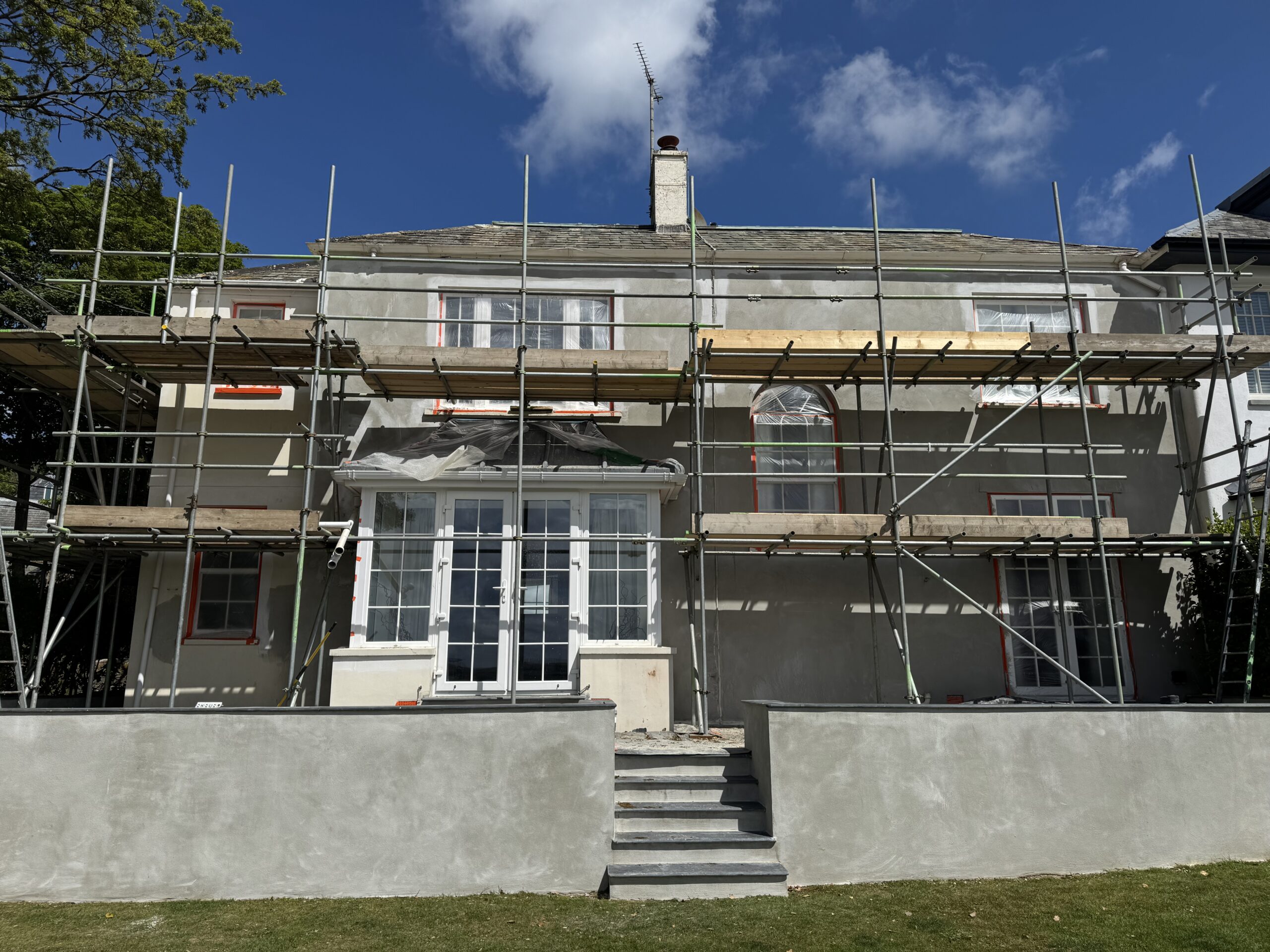 A front view of a two-story detached white house undergoing external rendering work while surrounded by construction scaffolding in Cornwall.