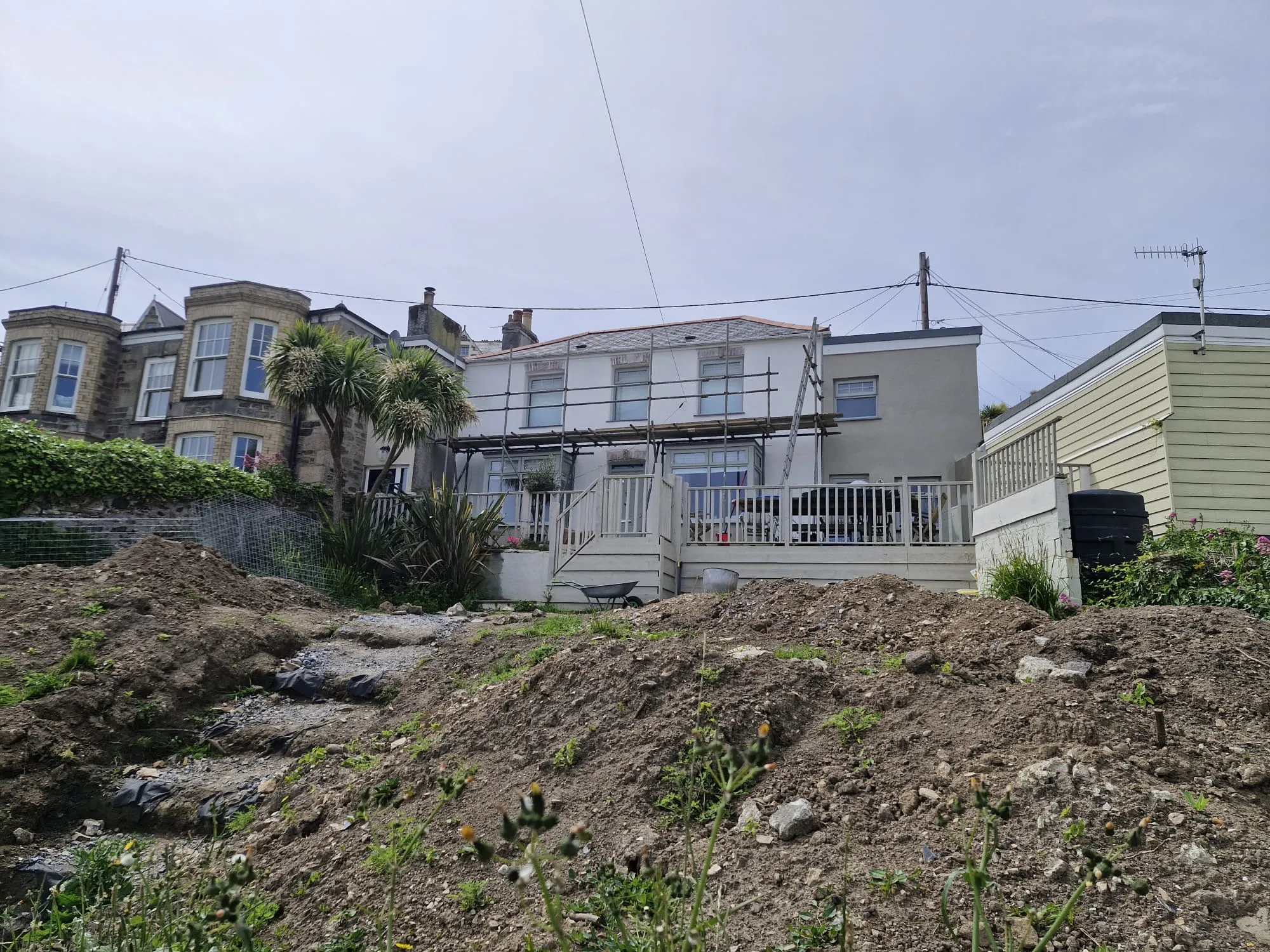An exterior view of a traditional stone house undergoing a rear extension with construction scaffolding setup against the building and groundworks excavation in Cornwall.